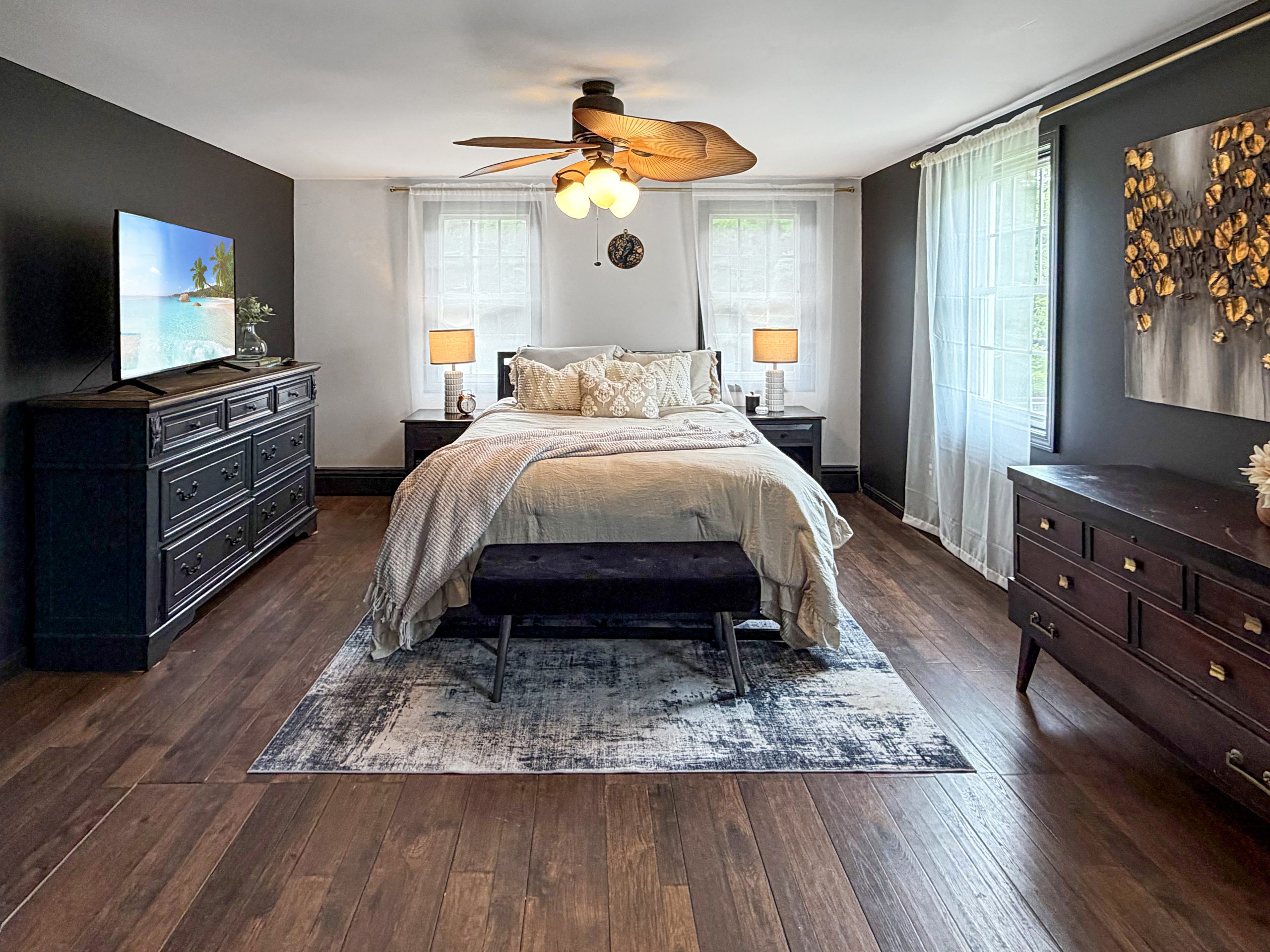 Staged primary bedroom with neutral bedding, dark wood furniture, layered rug, and warm lighting