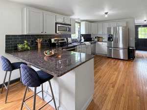 Bright kitchen with white cabinets, dark tile backsplash, granite counters, stainless steel appliances, and bar seating