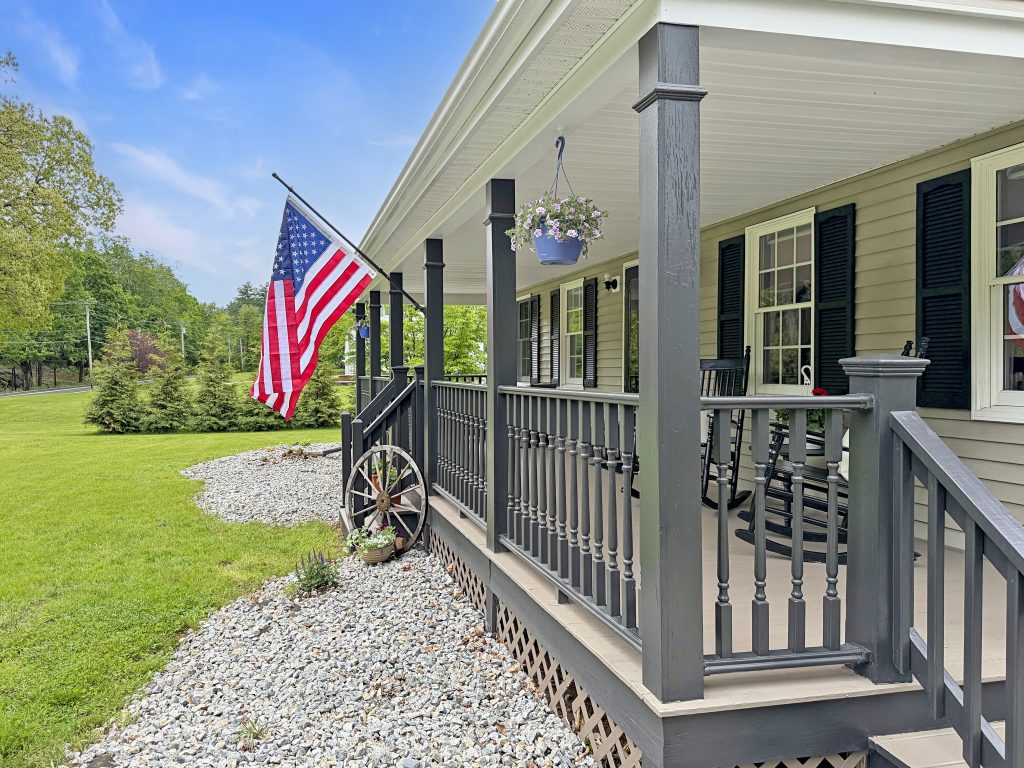 Welcoming farmhouse-style front porch with American flag, gray railings, black shutters, and landscaped yard